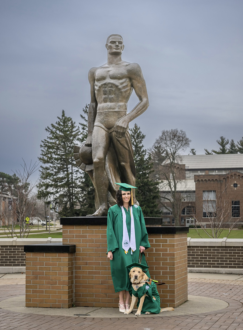 Alexandra Allers and her dog Leo post in front of the Spartan statue in commencement attire