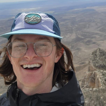 This photo of Colin Reese is a selfie taken outside. He is wearing glasses and a blue and white baseball cap.